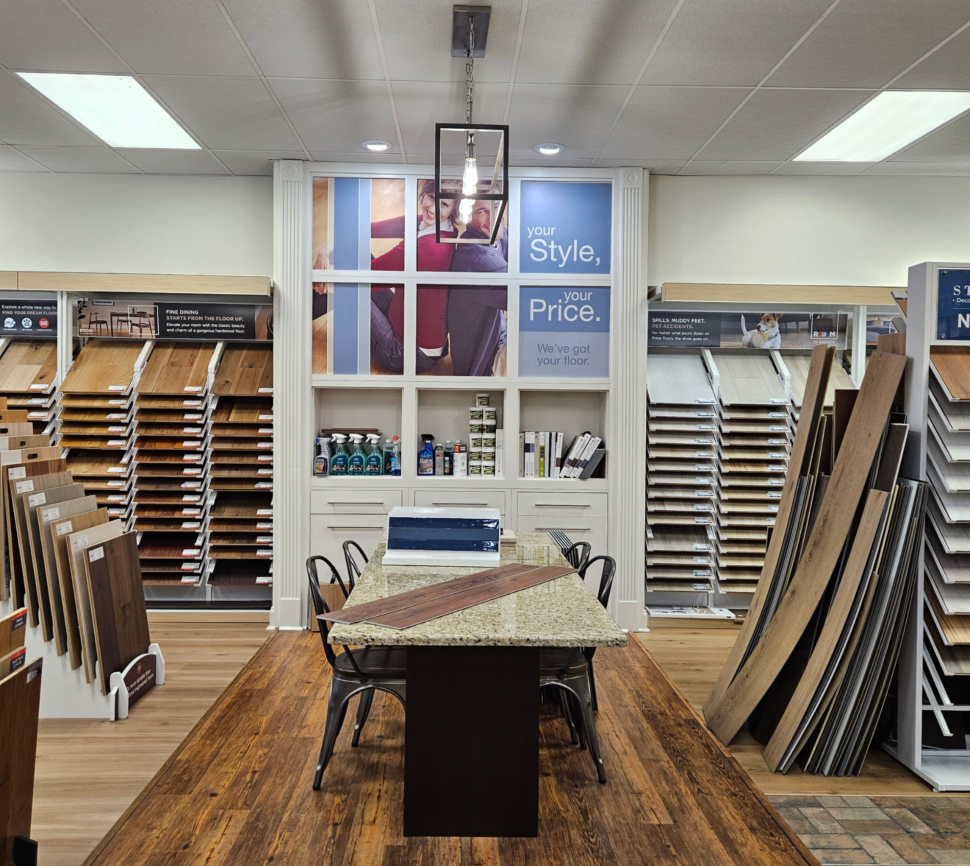 Interior of Noel Maestri's flooring showroom with rows of wood planks on both sides, a central display table, shelves of samples, and a banner reading “Style, Your Price.”