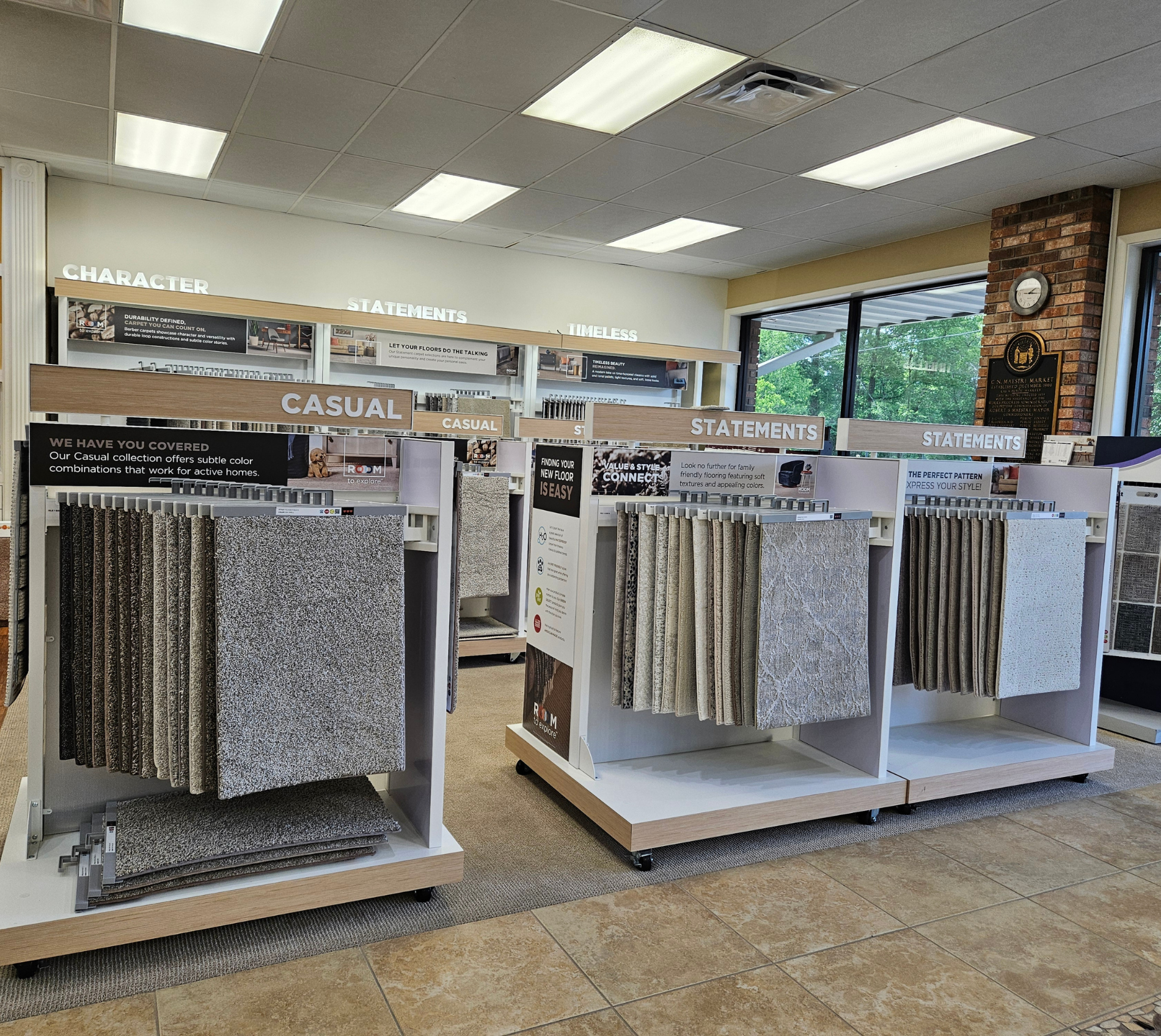 A flooring showroom with multiple carpet and tile sample displays on freestanding racks, labeled sections for casual and statements, and large windows along the back wall.