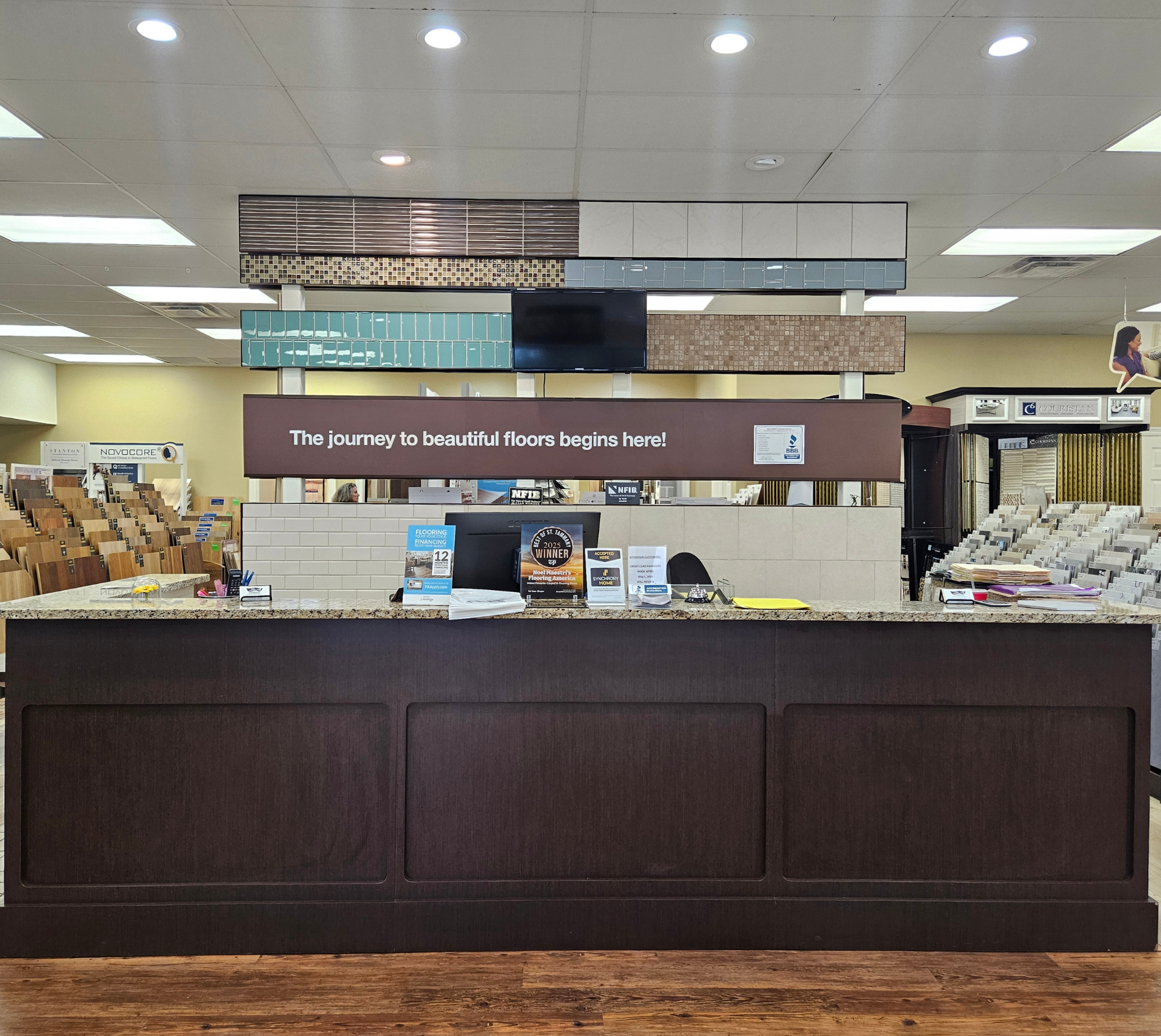 Flooring store front counter with displays and brochures, stacked samples in the background, and a banner reading “The journey to beautiful floors begins here!”